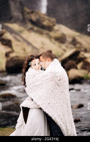 Un couple de mariage se tient sur le rivage d'une rivière de montagne sous une couverture de laine. Le marié épouse la mariée, essayant de la réchauffer. S Banque D'Images