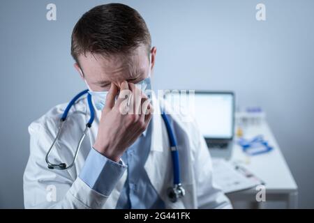 Jeune médecin européen allemand regardant l'appareil photo portant des lunettes et un masque dans le bureau de la clinique sur le lieu de travail avec le regard fatigué. Le sujet est Banque D'Images