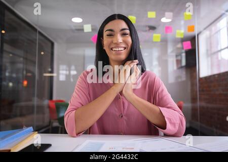Bonne femme d'affaires mixte de course assis à la table tout en ayant un appel vidéo souriant Banque D'Images