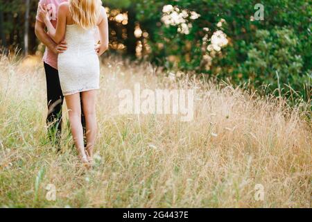 Un homme dans une chemise rose et une femme dans une robe blanche courte embrassent parmi les épillets, derrière eux sont des arbres verts Banque D'Images