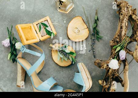 Les anneaux de mariage se trouvent sur un support en bois entouré de sandales avec talons, boîte en bois, parfum, fleurs et brindilles sur le sol Banque D'Images