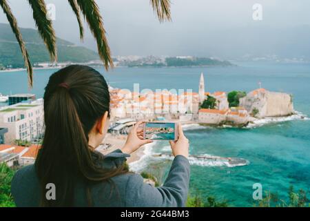 Une fille aux cheveux foncés prend des photos de la ville de Budva de loin sur smartphone Banque D'Images