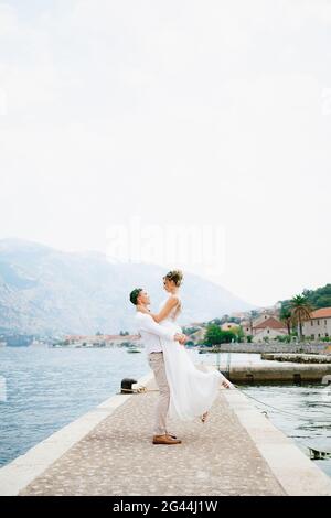 Le marié entoure la mariée dans ses bras sur le jetée dans la baie de Kotor près de la vieille confortable ville Banque D'Images
