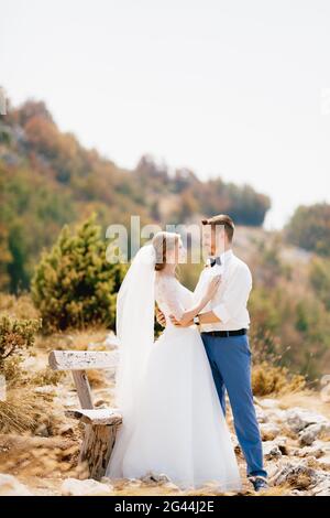 La mariée et le marié s'embrassent tendinement près d'un bois banc dans les montagnes et regarder l'un l'autre Banque D'Images