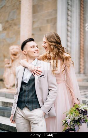 Mariée avec un bouquet de fleurs embrasse les épaules de groom souriant sur les marches à l'entrée de la basilique de Santa Maria Magg Banque D'Images