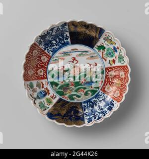 Soucoupe-plat avec parois et fleurs cannelées et coquillier au-dessus des vagues. Echelle de porcelaine avec paroi striée et bord de la bande, peinte en bleu sous-glaçure et sur le bleu de glaçure, rouge, vert, jaune, aubergine, noir et or. Sur l'appartement un médaillon avec des fleurs et des coquillages prunus au-dessus des vagues tourbillonnantes; le mur divisé en huit surfaces avec la même décoration de l'autre côté à l'autre côté: les boissons au lotus ont protsé sur un sol rouge, des arbres en fleurs avec oiseau et papillon, un paysage en or contre un bleu clair (poudre bleue) sol et les coûts (feuille d'artemisia) avec des symboles de bonheur (trépied Banque D'Images