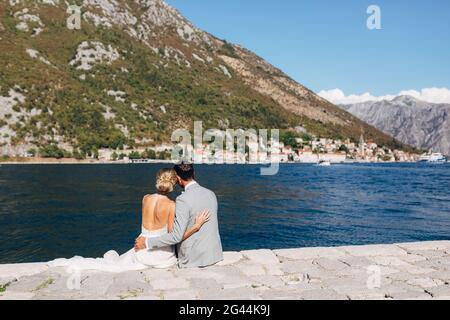La mariée et le marié sont assis embrassant sur la jetée dans la baie de Kotor, en face d'eux est la vieille ville Banque D'Images