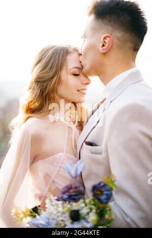 Le marié embrasse le front de la mariée souriante dans une belle robe rose avec un bouquet de fleurs Banque D'Images