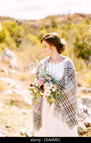 Mariée souriante avec un châle à carreaux sur ses épaules et un beau bouquet de fleurs dans ses mains sur un arrière-plan de la nature Banque D'Images