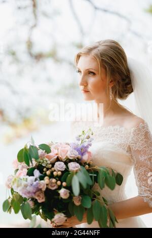 Une mariée sophistiquée se tient avec un bouquet de mariage dans ses mains sous les branches d'un arbre, gros plan Banque D'Images