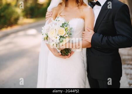 Le marié épouse les épaules de la mariée, qui tient un bouquet de mariage avec des fleurs blanches, gros plan Banque D'Images