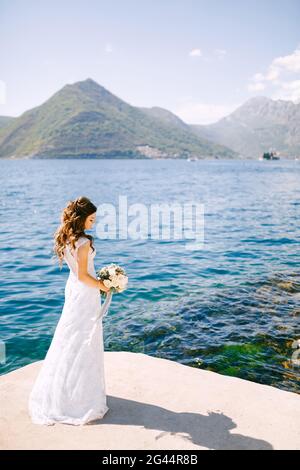 Une mariée élégante avec un bouquet dans ses mains se tient Sur un quai dans la baie de Kotor près de la eau bleue transparente Banque D'Images