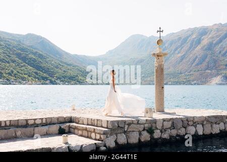 Une mariée marche le long d'un quai en pierre à côté d'une vieille colonne dans la baie de Kotor, sa jupe se dépare dans le vent Banque D'Images