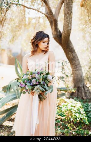 Jolie mariée dans une belle robe se tient pensivement contre un arrière-plan de l'arbre avec un magnifique bouquet de fleurs en elle mains Banque D'Images