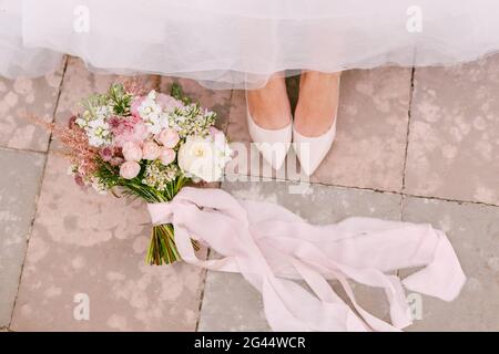 Les jambes de la mariée dans des chaussures blanches gracieux Peek hors de sous la jupe, à côté du bouquet de la mariée se trouve, gros plan Banque D'Images