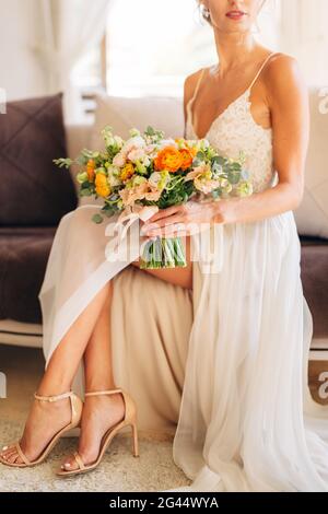 Une mariée habillée dans une robe de mariage et des sandales à talons hauts est assise sur un canapé dans la chambre et accueille un mariage bouquet entre ses mains Banque D'Images