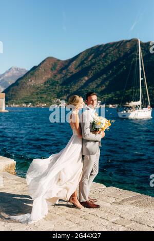 La mariée et le marié embrassent sur la jetée dans la baie de Kotor, derrière eux sont des montagnes vertes et un yacht blanc Banque D'Images