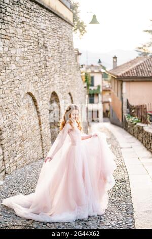 Mariée souriante dans une belle longue robe rose se dresse sur les pavés d'une vieille rue à Bergame, Italie Banque D'Images