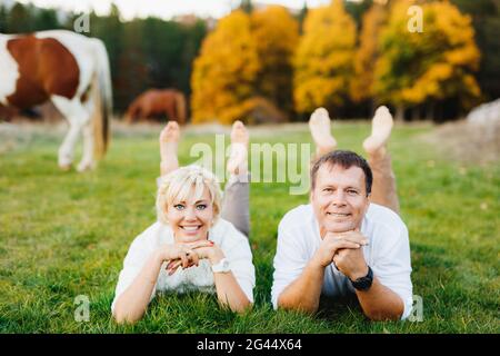 Le couple se trouve sur la pelouse avec leurs bras repliés sous leurs chins dans la forêt d'aurudes. Les chevaux se broutent en arrière-plan Banque D'Images