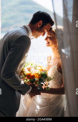 La mariée et le marié sont assis à la fenêtre s'embrassant, souriant et tenant les mains de l'autre. Dans leurs mains une brida Banque D'Images