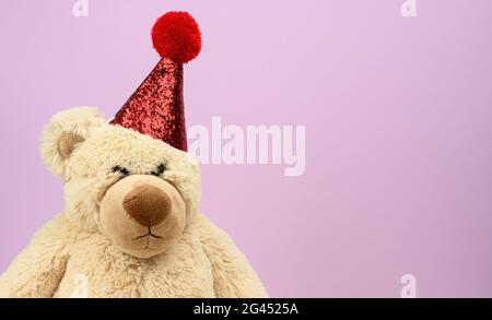 Un ours en peluche beige triste dans une casquette rouge se trouve sur un fond violet Banque D'Images