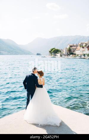 La mariée et le marié sont debout, embrassant et embrassant sur la jetée près de la vieille ville de Perast, vue arrière Banque D'Images