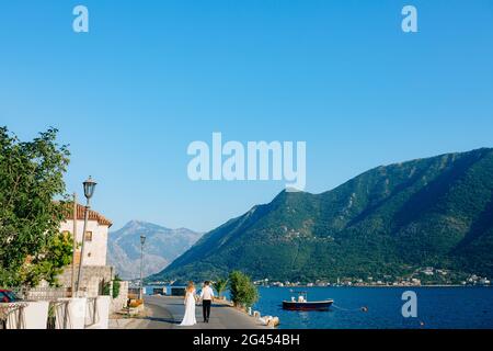 La mariée et le marié marchent en tenant les mains sur la jetée Dans Perast avec la toile de fond d'une vue pittoresque de La baie de Kotor Banque D'Images