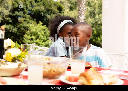 Une fille afro-américaine chuchote sur l'oreille de son frère lors d'un déjeuner familial dans le jardin Banque D'Images