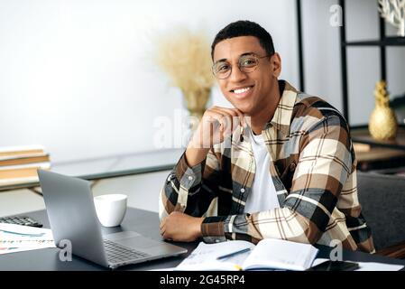 Portrait beau sûr de succès jeune course mixte latino homme, indépendant ou directeur, portant une chemise et des lunettes, assis au bureau avec ordinateur portable, regardant l'appareil photo et souriant amical Banque D'Images