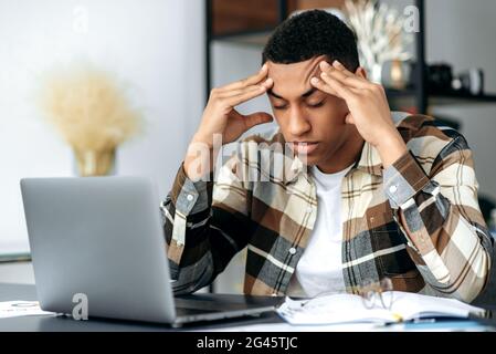 Déprimé latino jeune homme, dans des vêtements décontractés et élégants, indépendant, assis à son bureau, massant ses temples avec les yeux fermés, pensant à un projet, avoir un mal de tête, migraine, travail excessif Banque D'Images