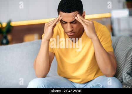 Frustré triste hispanique gars dans un t-shirt jaune assis sur un canapé à la maison, moasque ses temples avec ses yeux fermés, il a des problèmes personnels, maux de tête, migraine, stress, pense à résoudre le problème Banque D'Images