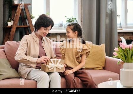 Bonne grand-mère ouvrant la boîte-cadeau tout en parlant à la petite-fille dans la salle de séjour Banque D'Images