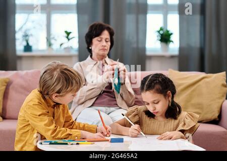 Deux enfants dessinant à la table tandis que leur grand-mère tricoter sur le canapé Banque D'Images