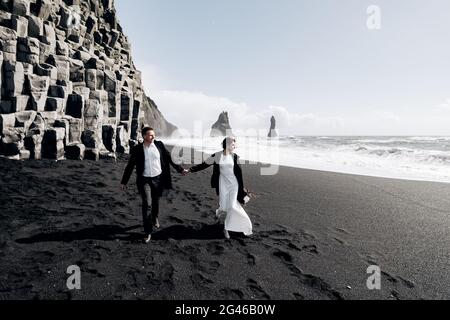 Destination mariage Islande. Un couple de mariage marche le long de la plage de sable noir de Vik, près de la roche de basalte, sous forme de pil Banque D'Images