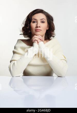 Belle femme brune habillée dans un grand chandail blanc en laine est assise à table blanche. Banque D'Images