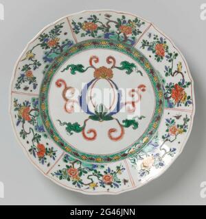 Assiette avec décoration à volutes foliaires et plantes à fleurs. Plaque à large bord de porcelaine, peinte en bleu sous-glaçure et sur le bleu glaçure, rouge, vert, jaune, aubergine, noir et or. Sur l'appartement une cartouche vide décorée de radeau de feuilles; le mur avec un motif géométrique contre un sol tacheté vert; le bord avec une décoration commerciale remplie de plantes à fleurs, d'oiseaux et d'insectes; le dos avec trois branches de fleurs. Sur le dessous étiqueté avec un brûleur à encens dans un double cercle. La plaque a été cassée. Famille verte. Banque D'Images