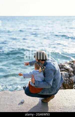 Papa s'est accroupi en tenant la petite fille sur ses genoux sur un grand rocher. Papa pointe son doigt sur la mer vers sa fille. Retour Banque D'Images