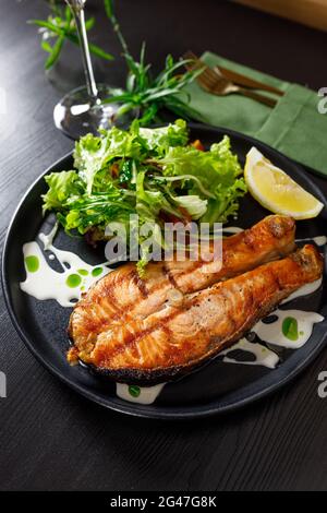 Un morceau de saumon grillé poivre citron et sel sur une assiette noire avec des feuilles de laitue sur fond de pierre Banque D'Images