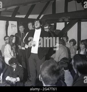 1970, musique folklorique historique, dans une salle d'un pub de campagne avec poutres en bois, un public écoutant un chanteur de folk mâle barbu chantant une chanson au Castle Inn, Colchester, Essex, Angleterre, Royaume-Uni. Banque D'Images