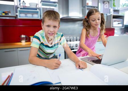 Garçon faisant ses devoirs pendant que fille utilisant un ordinateur portable dans la cuisine Banque D'Images
