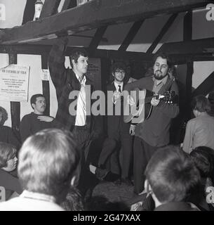 1970, historique, musique folk, dans un pub de campagne avec poutres en bois, un public écoutant un homme dans un gilet debout, jouant une guitare, au Castle Inn, Colchester, Essex, Angleterre, Royaume-Uni. Affiche publicitaire sur le mur, faisant la promotion de « une soirée de folk » avec Shibase & Dolly Collins, Dave & Toni Arthur, Bob & Carol Pegg et Dave & Dave au Stevenage College of Elder Education. Banque D'Images