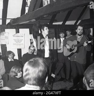 1970, historique, musique folk, à l'intérieur d'une salle à poutres apparentes dans un pub, un public de jeunes écoutant un musican folklorique, un homme dans un gilet jouant une guitare, The Castle Inn, Ongar Rd, Brentwood, Essex, Angleterre, Royaume-Uni. Affiche publicitaire sur le mur, indiquant « une soirée de folk » avec des sœurs, Shirley & Dolly Collins, Dave & Toni Arthur, Bob & Carol Pegg, et Dave & Dave au Stevenage College of Elder Education, promu par le Red Lion Folk Club. Banque D'Images