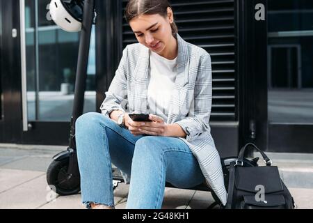 Femme d'affaires tapant sur un téléphone cellulaire tout en étant assise sur un scooter électrique poussée dans le bâtiment Banque D'Images