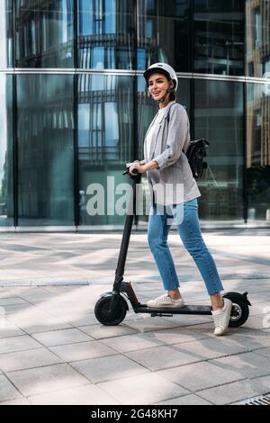 Jeune femme heureuse en mode décontracté au volant d'un scooter électrique. Femme d'affaires en casque de vélo debout sur e-scooter. Banque D'Images