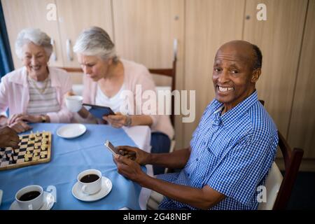 Homme âgé souriant utilisant un téléphone portable tout en prenant le thé avec des amis Banque D'Images