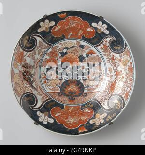 Plat avec un vase à fleurs, Feng Huangs, Shishi et oiseaux. Plat de porcelaine, peint en bleu sous-brillant et sur le rouge, noir et or brillant. Sur l'appartement un double médaillon avec un vase de fleur avec des chrysanthèmes et des pivoines. Le mur et le bord sont divisés en quatre compartiments avec deux shishi (chien lion) dans un paysage, deux oiseaux sur une branche d'un prunus et deux fois une cartouche en forme de ruyi avec un fenghuang sur un fond bleu avec des boissons florales. Le plat est pris dans un cadre métallique. Probablement un européen (français?) Imitation de l'Imari japonais. Banque D'Images