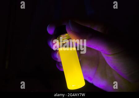 Femme chercheuse tenant une réaction photochimique jaune dans un flacon de verre sous lumière UV dans un laboratoire de chimie sombre pour la recherche biomédicale Banque D'Images