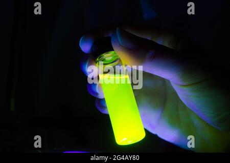 Femme chercheuse tenant une réaction photochimique verte dans un flacon de verre sous lumière UV dans un laboratoire de chimie sombre pour la recherche biomédicale Banque D'Images