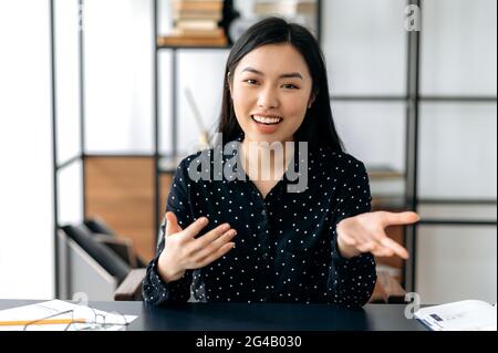 Communication vidéo en ligne. Sympathique, jolie, intelligente et confiante jeune femme asiatique, indépendante ou gestionnaire, assis au bureau, parler avec des collègues ou des amis par liaison vidéo, gestuelle des mains, souriant Banque D'Images