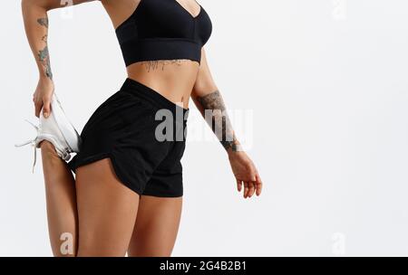 Vue en coupe basse d'une jeune femme en forme qui s'étire la jambe avant une course dans un studio de fitness, fond blanc. Coupe courte du bas du corps de l'athlète de fitness Banque D'Images